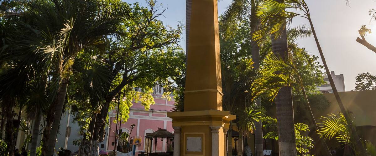 A tall monument surrounded by palm trees and a pink building in the background.