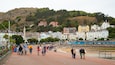 A coastal town with colorful buildings, a promenade, and a hill in the background.
