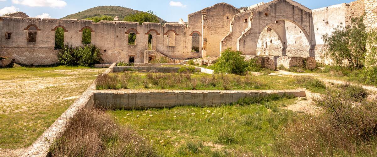 An ancient stone structure with arches and ruins, surrounded by grassy fields and a hill in the background.