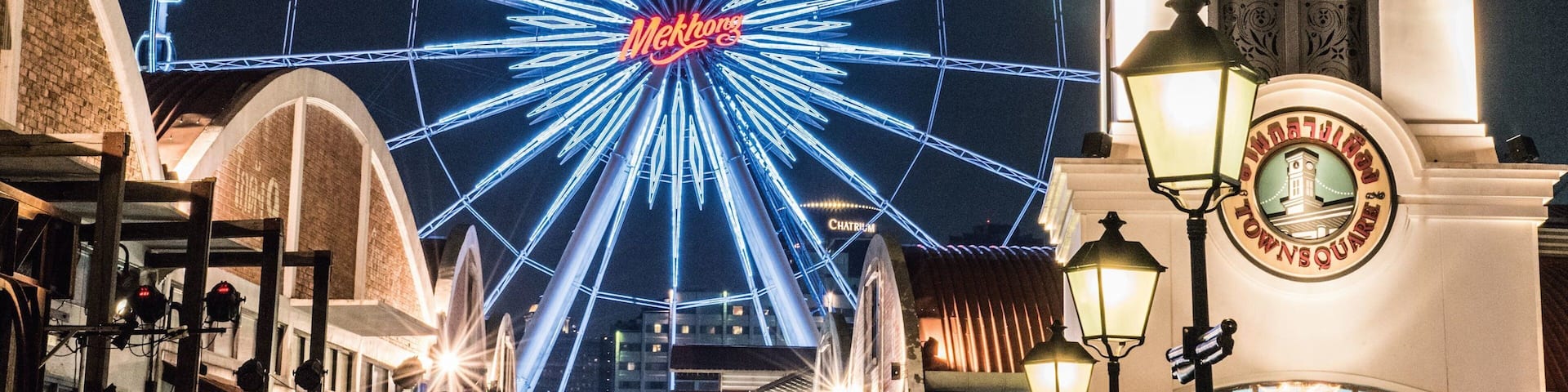 A brightly lit Ferris wheel with the word "Mekong" at its center, a clock tower with "TOWN SQUARE" inscribed, and a fountain in the foreground.