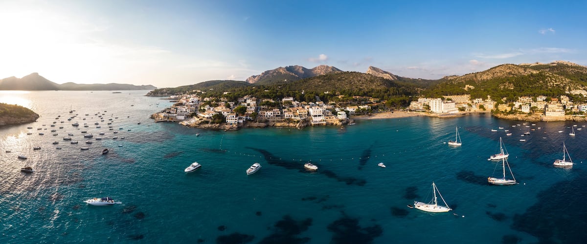 A coastal town with boats, clear blue waters, and mountains in the background.