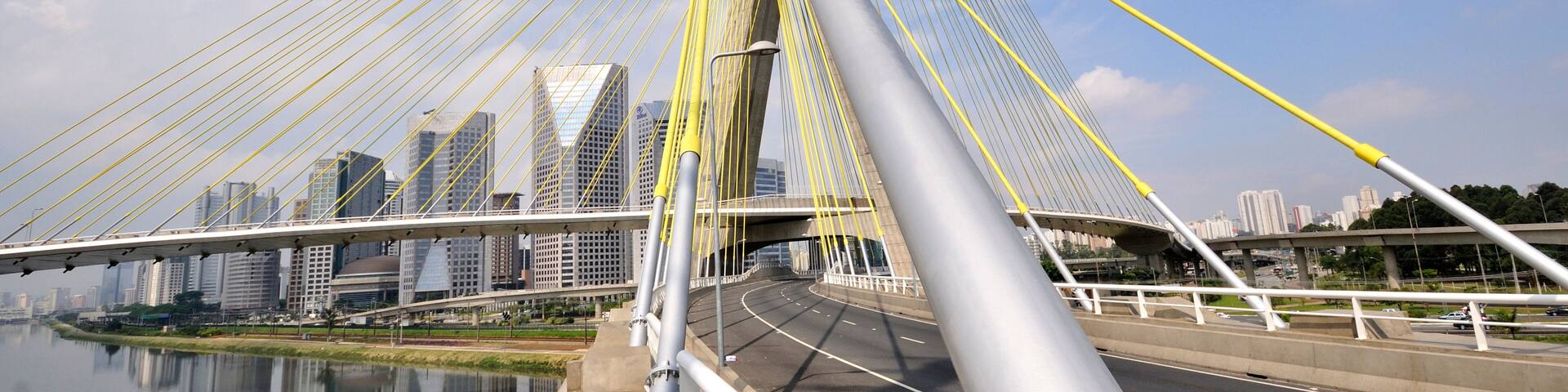 A cable-stayed bridge with yellow cables, a river below, and a city skyline in the background.