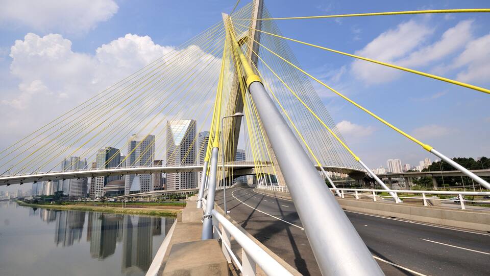 A cable-stayed bridge with yellow cables, a river below, and a city skyline in the background.