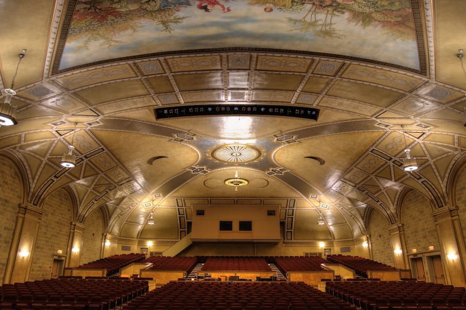 Interior de teatro ornamentado con un gran mural, techos abovedados y filas de asientos rojos.