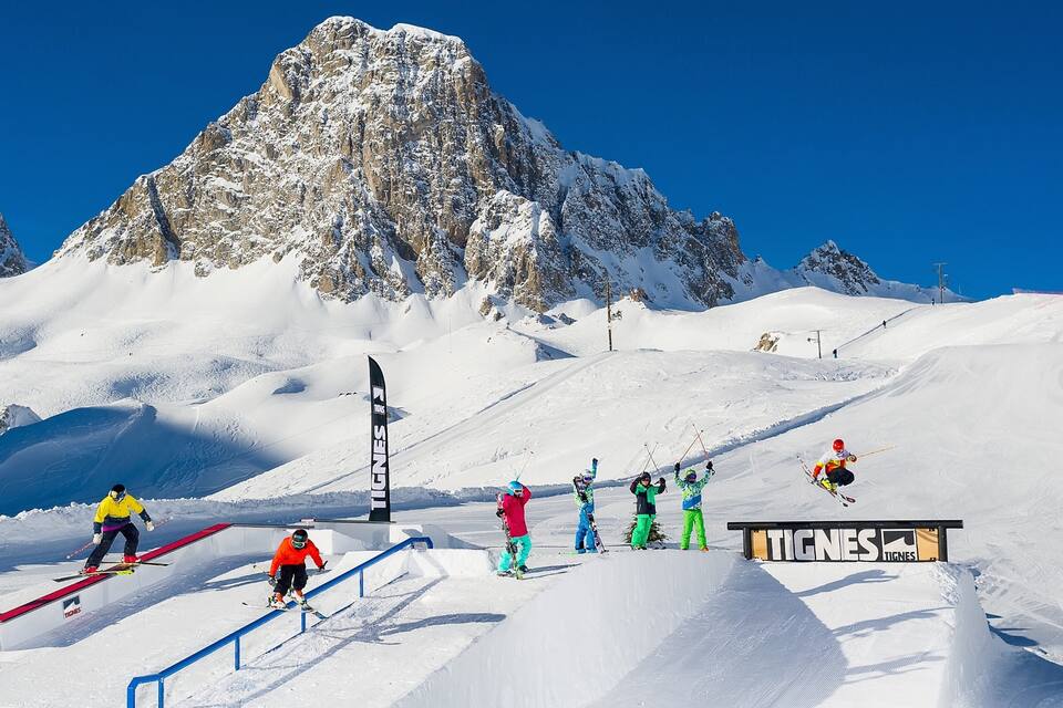 Een skipiste met een berg op de achtergrond, een Tignes bord en skiërs die trucjes uitvoeren.