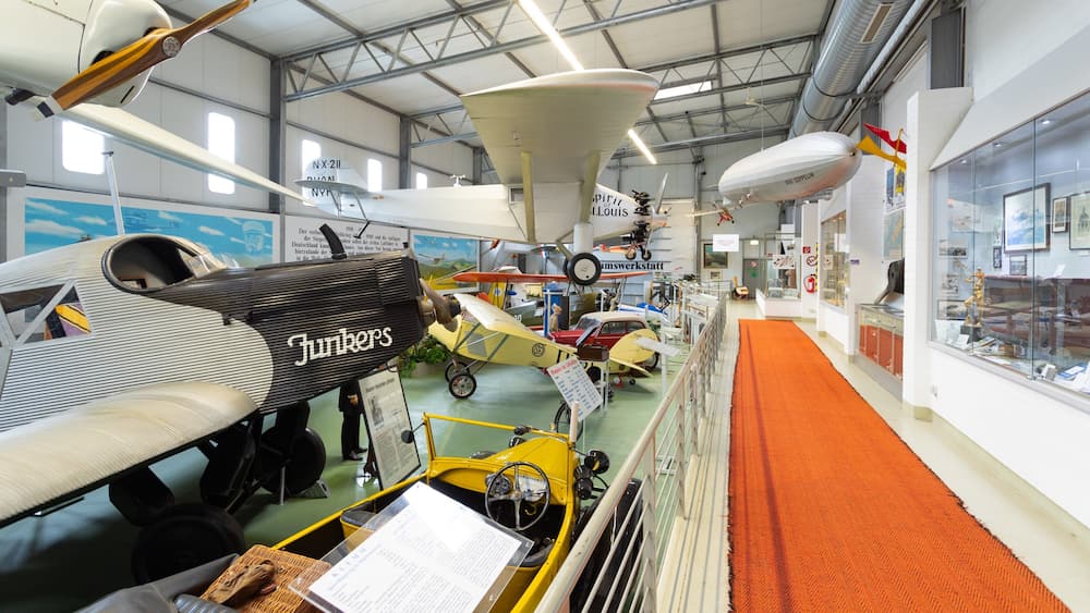 A museum with various aircraft on display, including a yellow car-shaped exhibit and a white biplane labeled 'Junkers'.