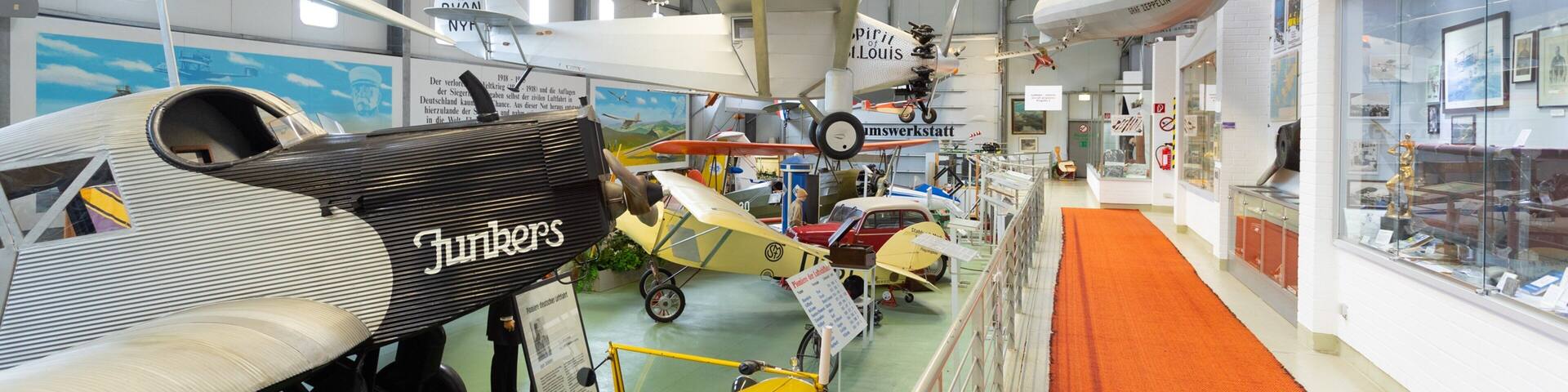 A museum with various aircraft on display, including a yellow car-shaped exhibit and a white biplane labeled 'Junkers'.