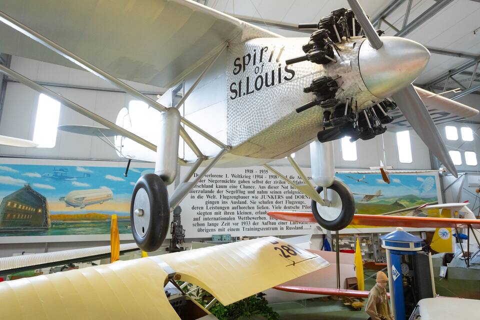 A vintage aircraft with 'Spirit of St. Louis' written on its side, displayed in a museum with other historical planes.