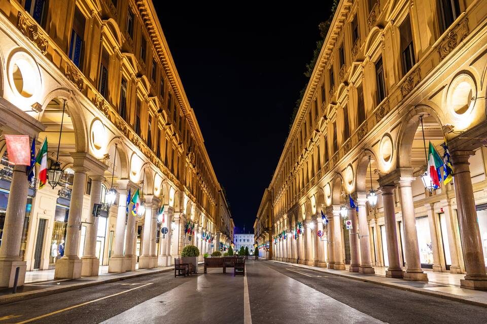 A nighttime view of a grand, illuminated street with classical architecture and multiple national flags.