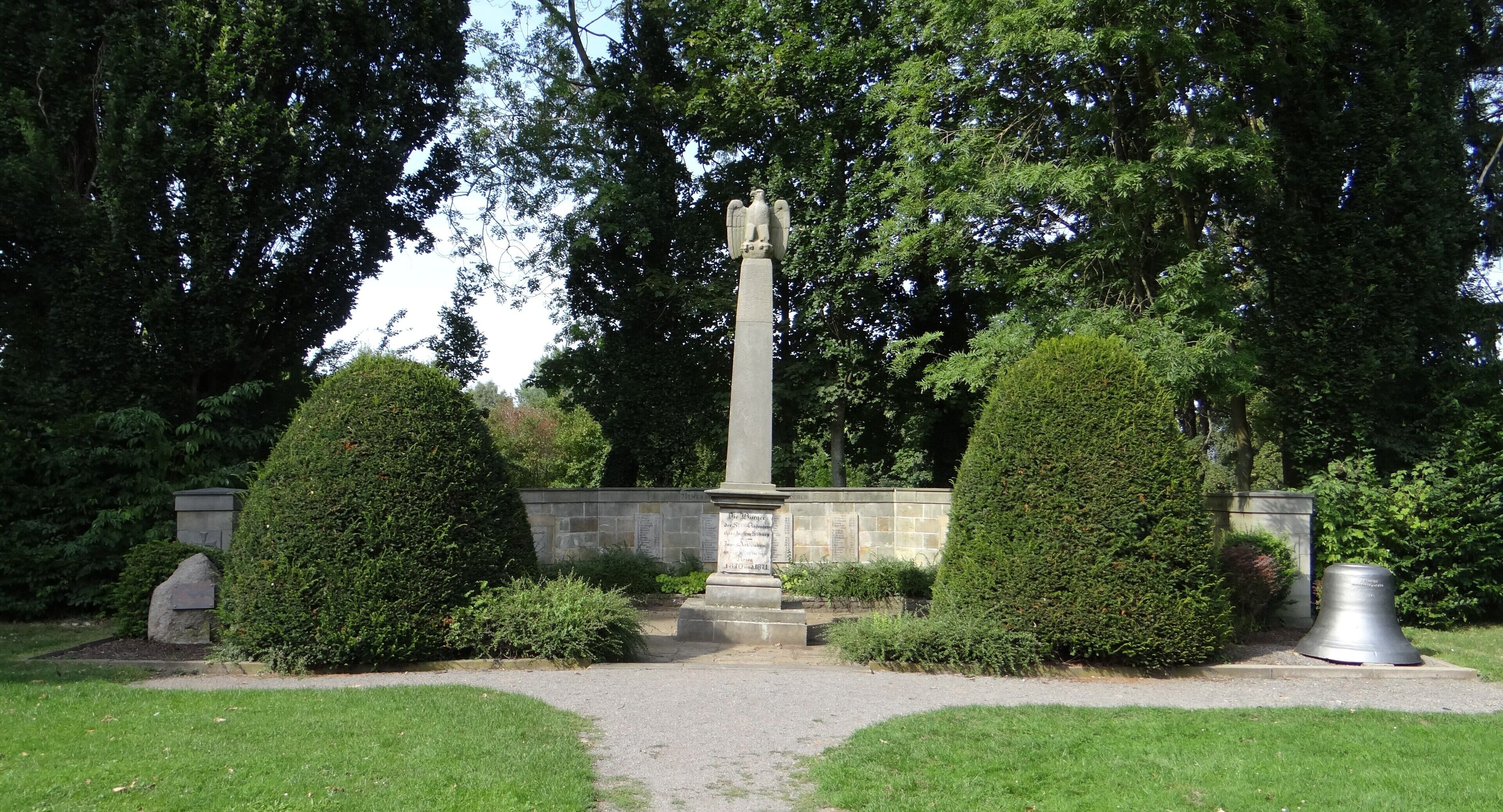 A memorial with an angel statue, surrounded by neatly trimmed hedges and a stone wall.