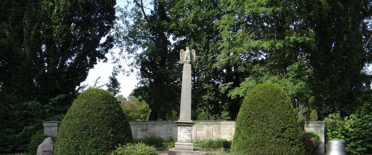 A memorial with an angel statue, surrounded by neatly trimmed hedges and a stone wall.