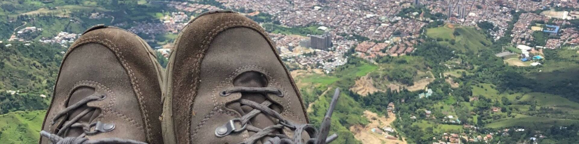 A pair of hiking boots with laces, a person wearing black pants, and a view of a cityscape with mountains in the background.