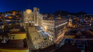 A historic cathedral with a prominent bell tower, surrounded by a densely populated urban area with illuminated buildings at night.