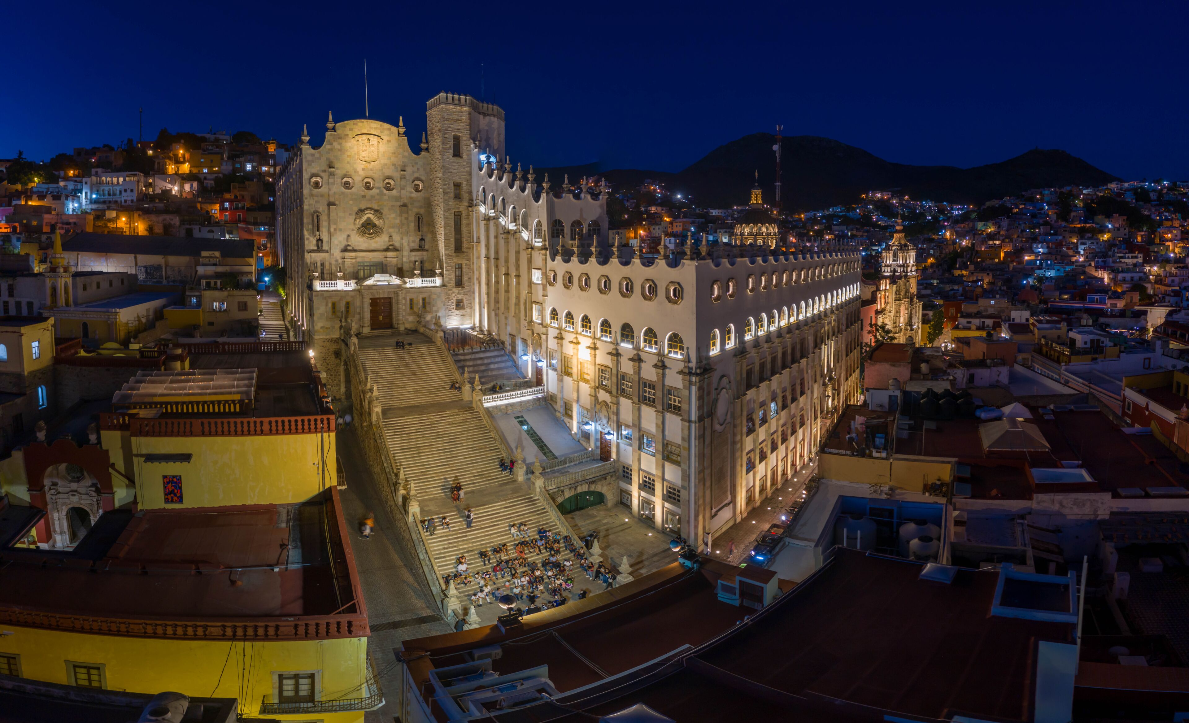 A historic cathedral with a prominent bell tower, surrounded by a densely populated urban area with illuminated buildings at night.