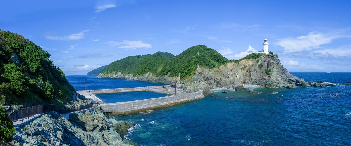A coastal landscape with a lighthouse on a rocky island, a clear blue sky, and calm sea.