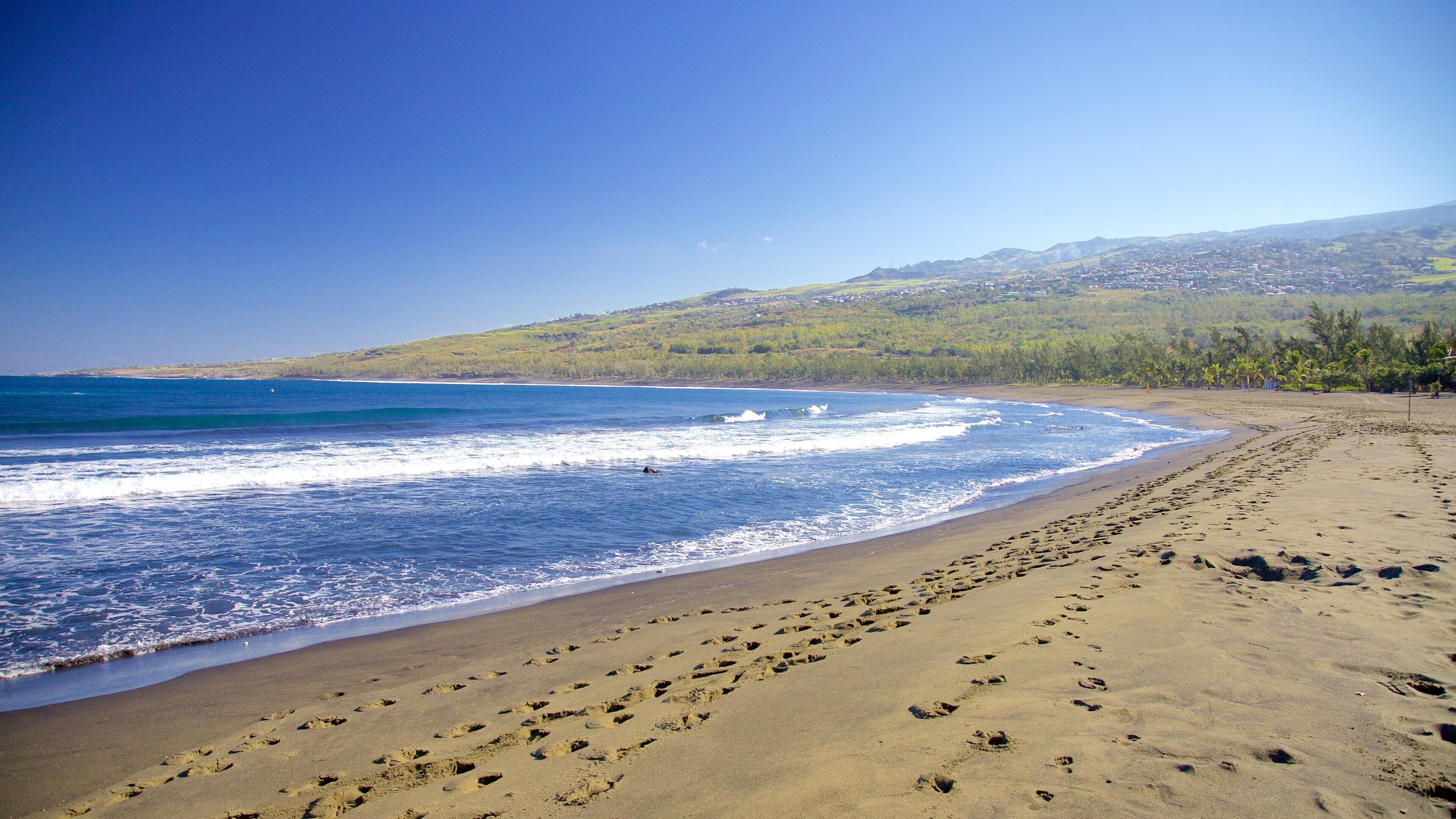 A sandy beach with footprints leading into the ocean, waves crashing onto the shore, and a hilly landscape in the background.