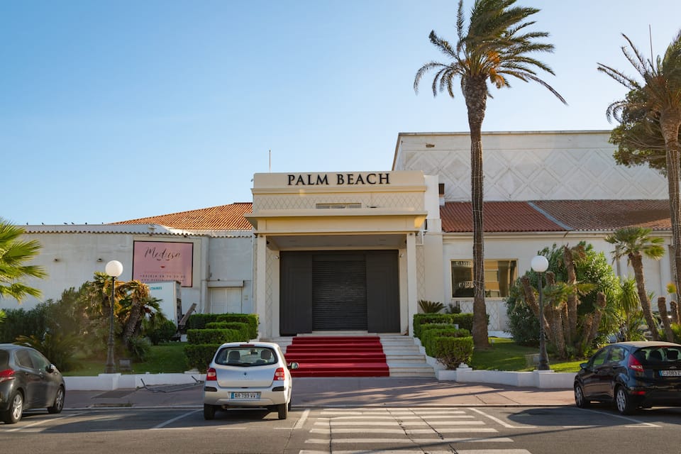 Een gebouw genaamd "Palm Beach", met een bord, palmbomen en geparkeerde auto's.