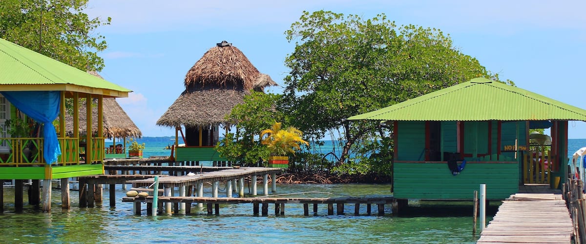 A wooden pier leading to overwater bungalows with thatched roofs, surrounded by clear blue water and lush greenery.