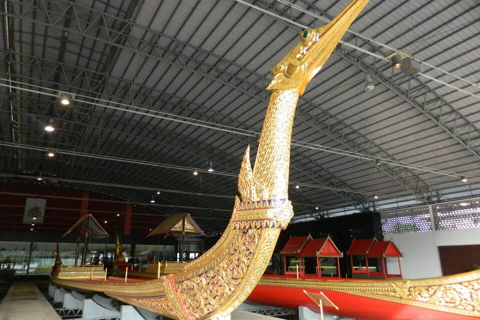 A large, ornate boat with intricate golden details and a pointed prow, displayed indoors with a high ceiling and industrial lighting.