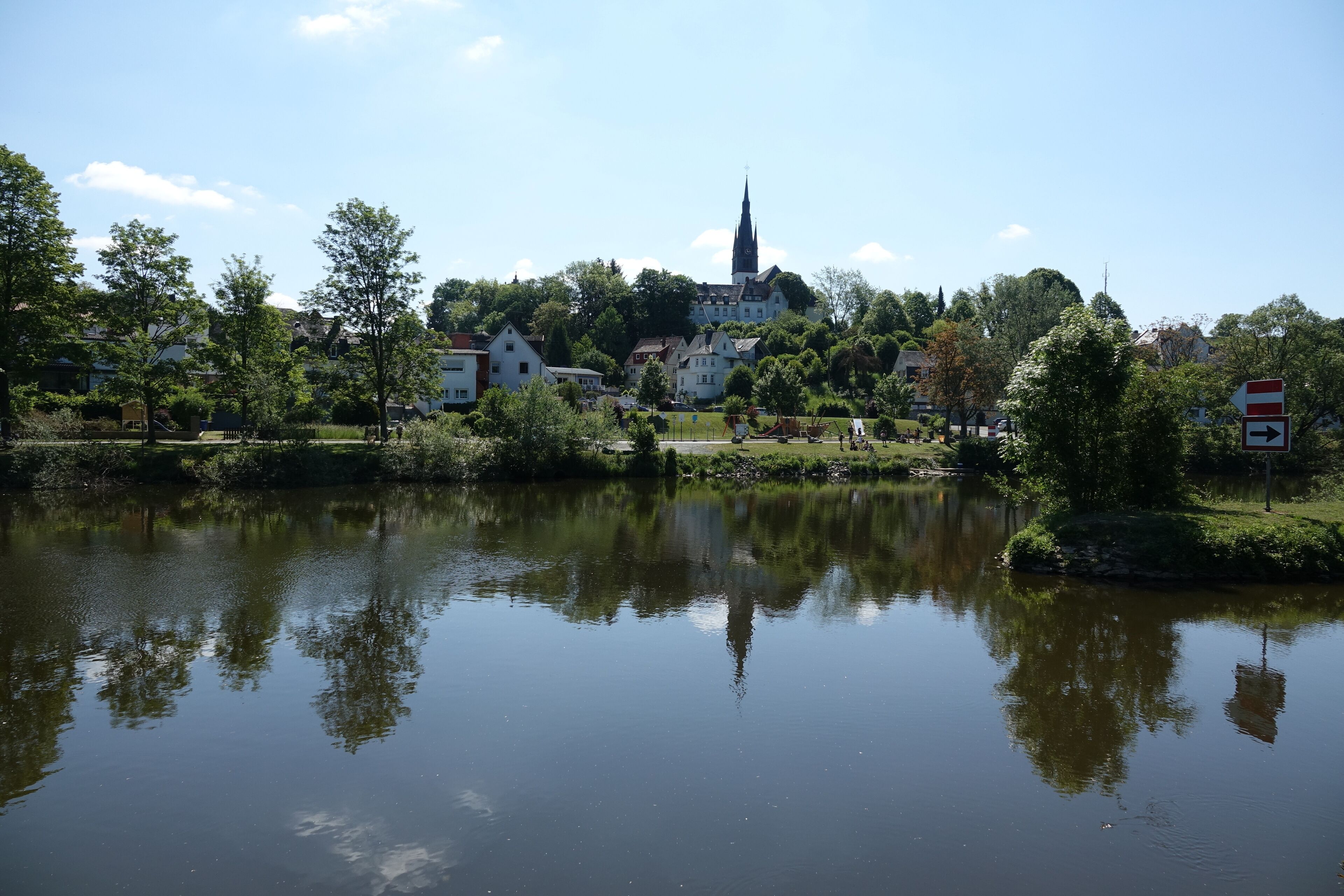 A calm lake with reflections of trees and buildings, surrounded by greenery and a church spire in the distance.