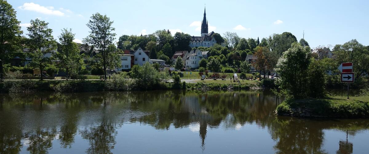 A calm lake with reflections of trees and buildings, surrounded by greenery and a church spire in the distance.