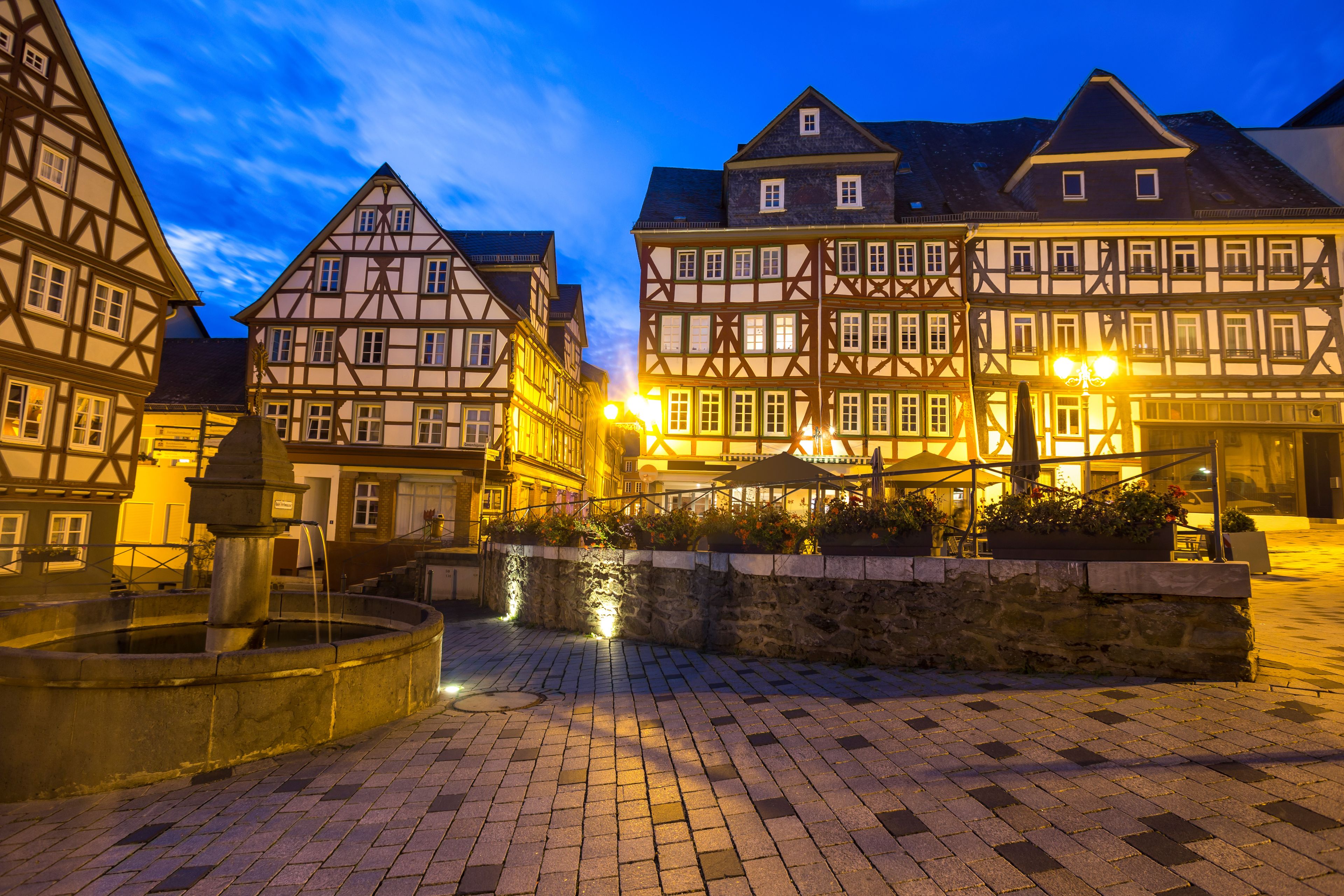A historic European town square at dusk with illuminated half-timbered buildings and a fountain.