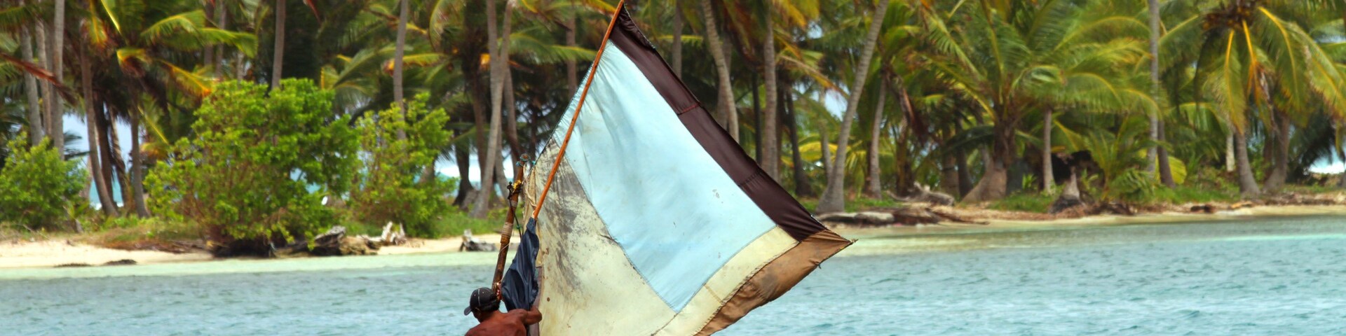 Un hombre en una canoa con vela en un mar turquesa, rodeado de palmeras.