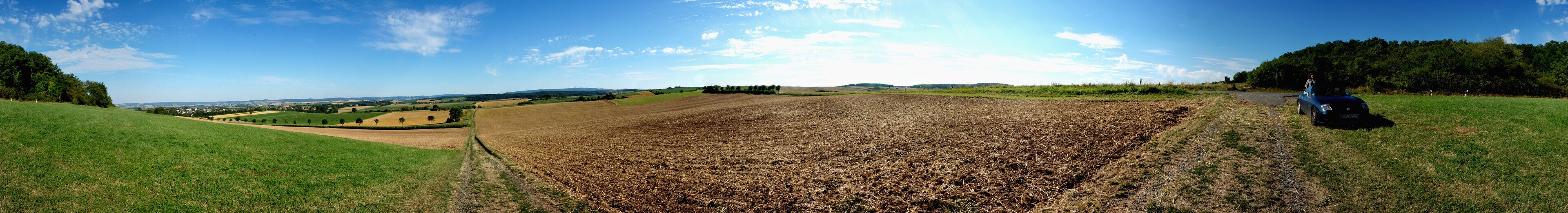 A rural landscape with fields, a dirt path, and a car parked on the side.