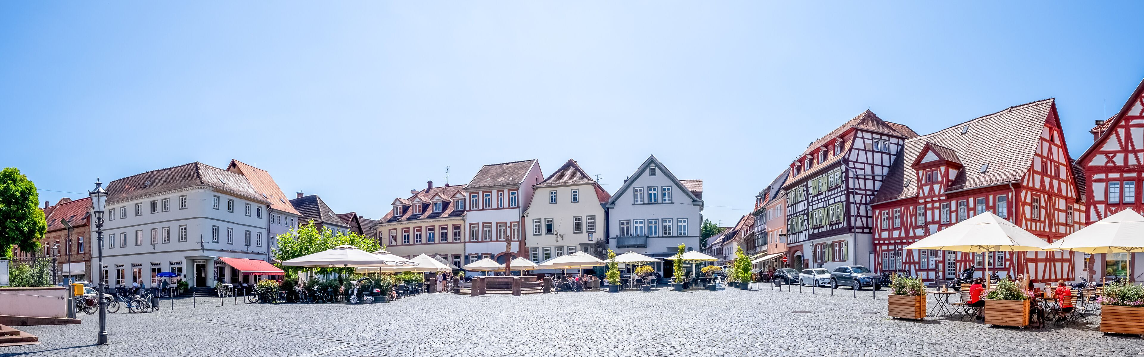 A cobblestone square with traditional European architecture, outdoor cafes, and parked cars.
