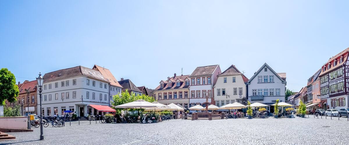 A cobblestone square with traditional European architecture, outdoor cafes, and parked cars.