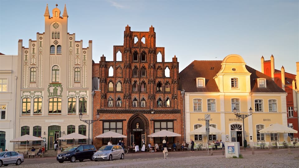 Uma praça histórica de cidade europeia, com uma mistura de arquitetura gótica e renascentista, incluindo uma igreja imponente com torre de relógio e um edifício tradicional de tijolos com janelas em arco.