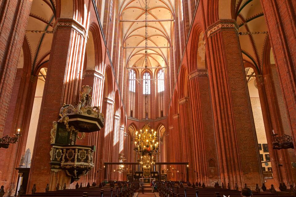 A cathedral interior with high vaulted ceilings, red brick walls, and a large chandelier.