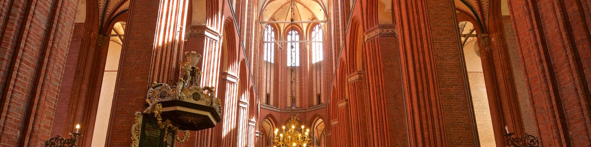 Interior de una catedral con altos techos abovedados, paredes de ladrillo rojo y una gran lámpara de araña.