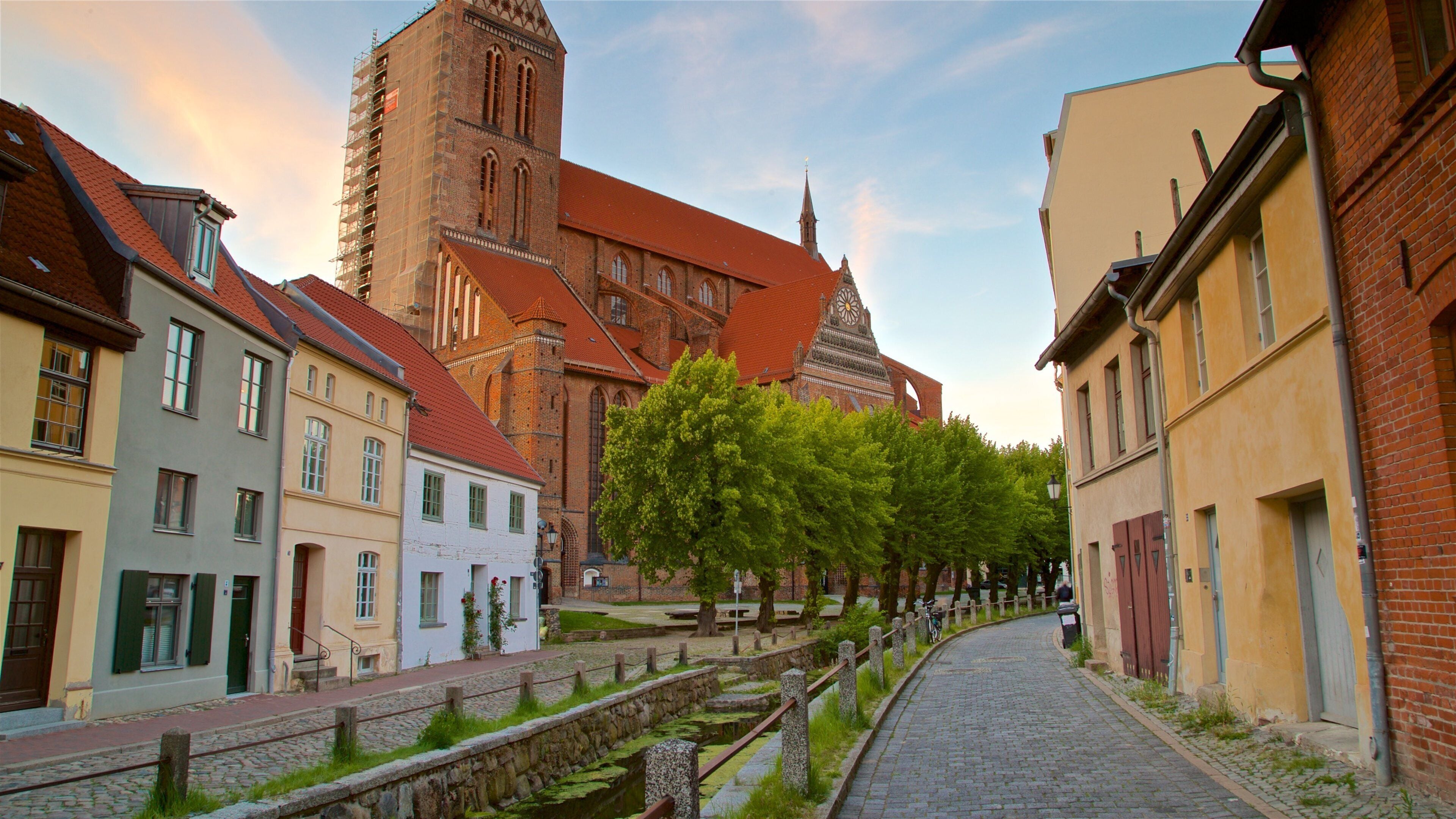 Eine Kopfsteinpflasterstraße säumen historische Gebäude, eine Kirche mit hohem Turm und ein Kanal mit einer Steinbrücke.