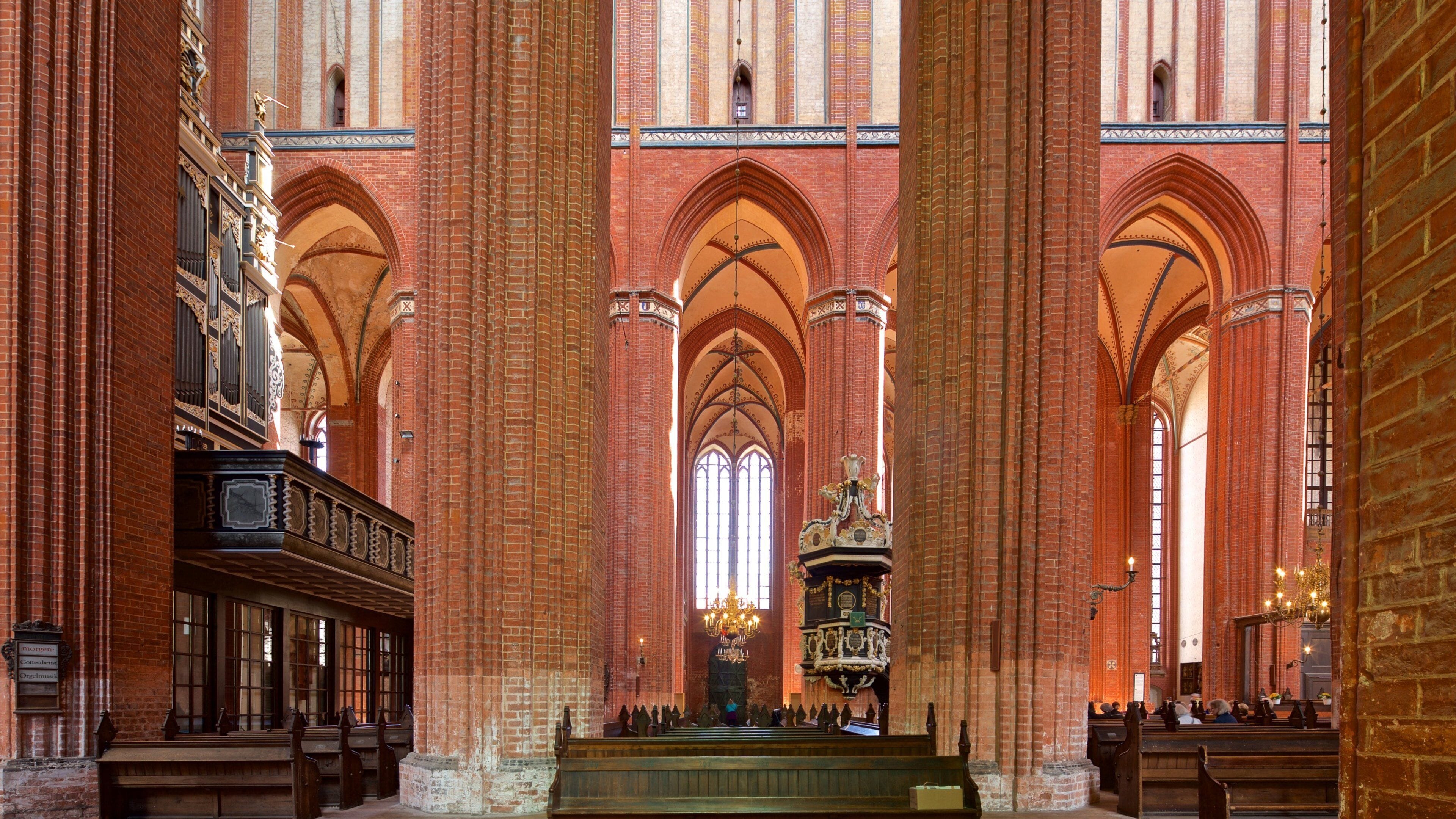 L’intérieur d’une cathédrale, avec de hauts plafonds en voûte, des murs en briques rouges et des bancs en bois richement décorés.