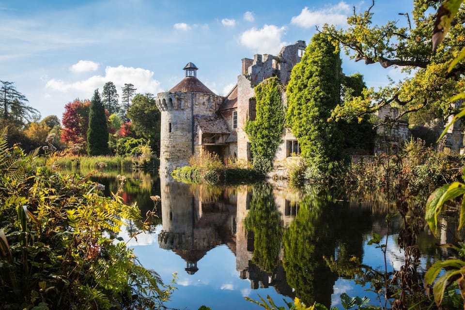 A historic castle with a moat, surrounded by lush greenery and a clear reflection in the water.