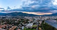 Een panoramisch uitzicht op een kuststad met een rivier, bergen op de achtergrond en een combinatie van woon- en bedrijfspanden.