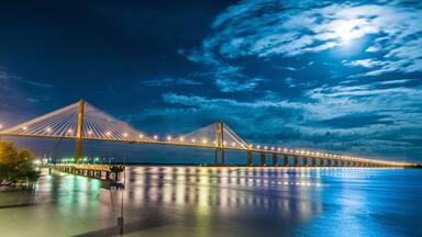 A nighttime view of a long suspension bridge with illuminated cables, spanning over a calm body of water.