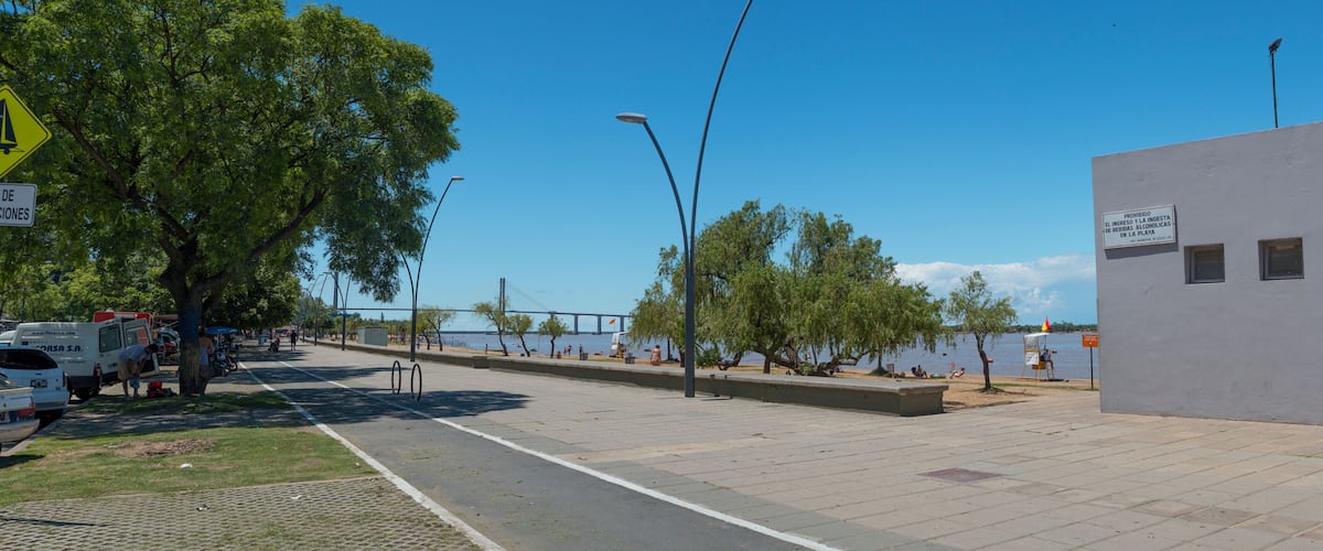 A waterfront promenade with a white building, street lamps, and a bridge in the background.