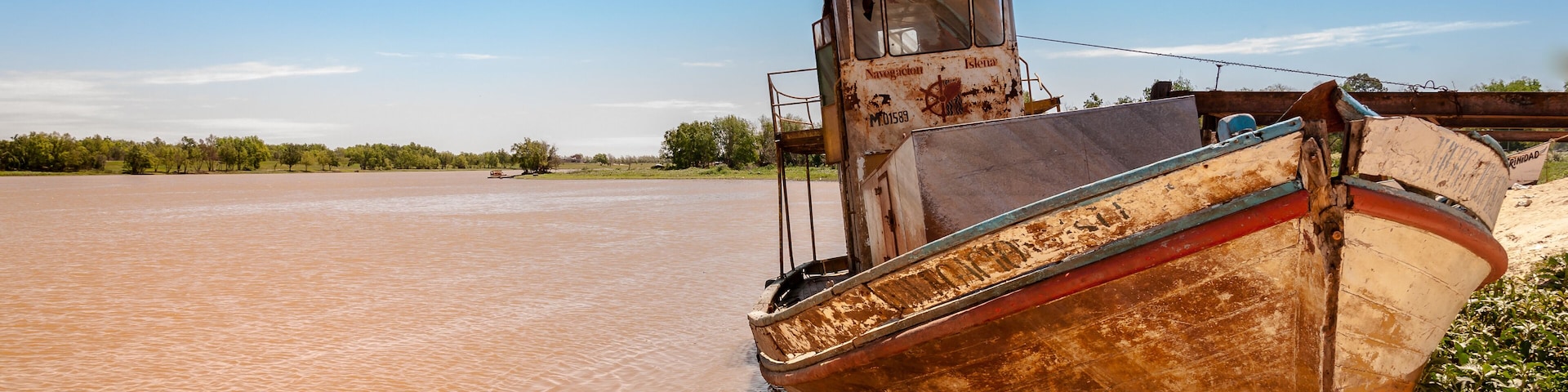 Un bote oxidado sobre una orilla arenosa con un río embarrado al fondo.