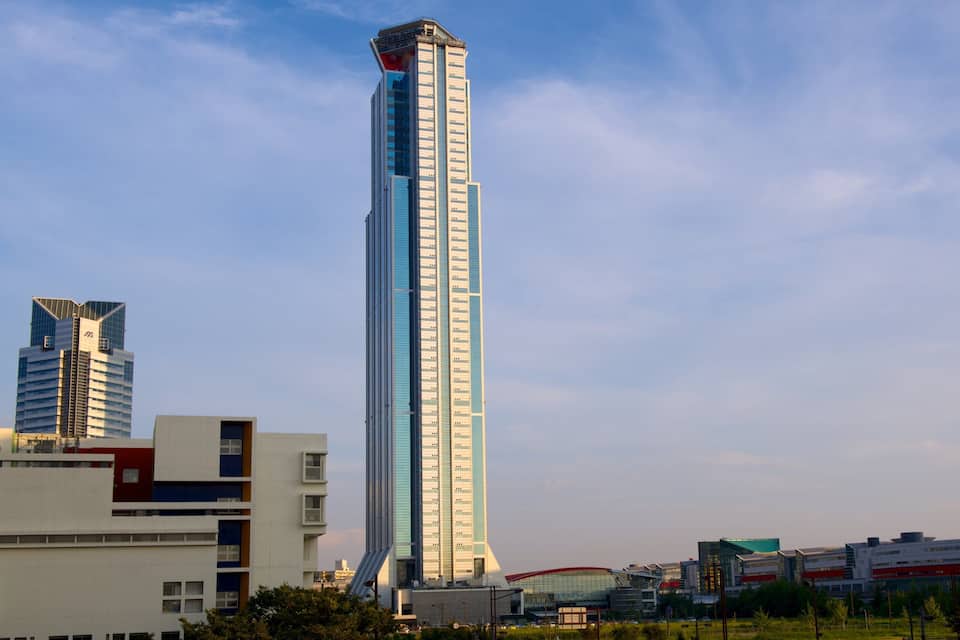 A tall skyscraper with a unique top design, surrounded by modern buildings and a clear sky.