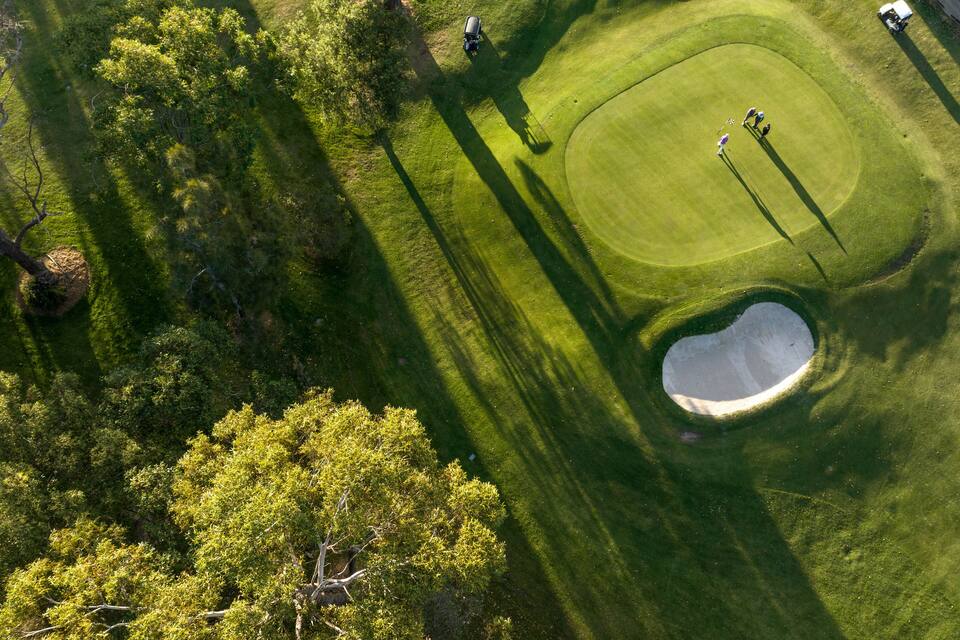 An aerial view of a golf course with a sand bunker, trees, and players.