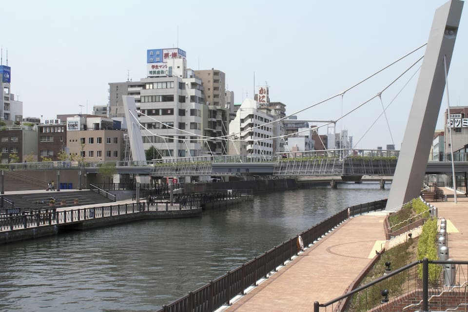 A riverside walkway with a modern bridge and buildings in the background.