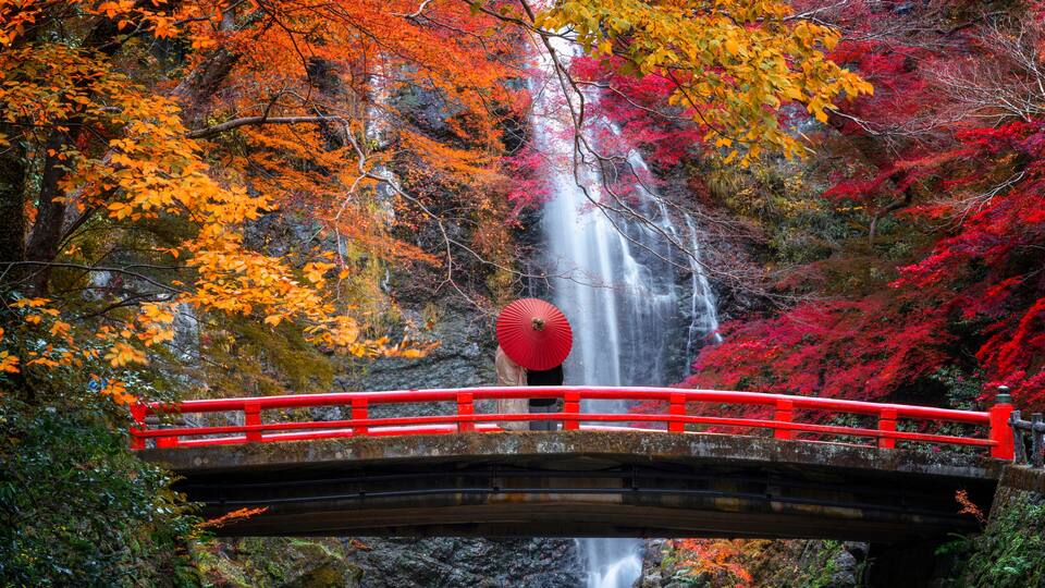 A red bridge over a waterfall with autumn foliage in the background.