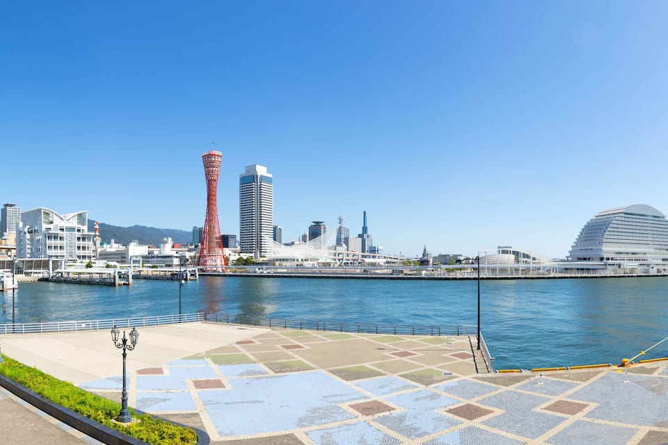 A waterfront cityscape with a modern tower, a distinctive red tower, and a large cruise ship docked at the pier.