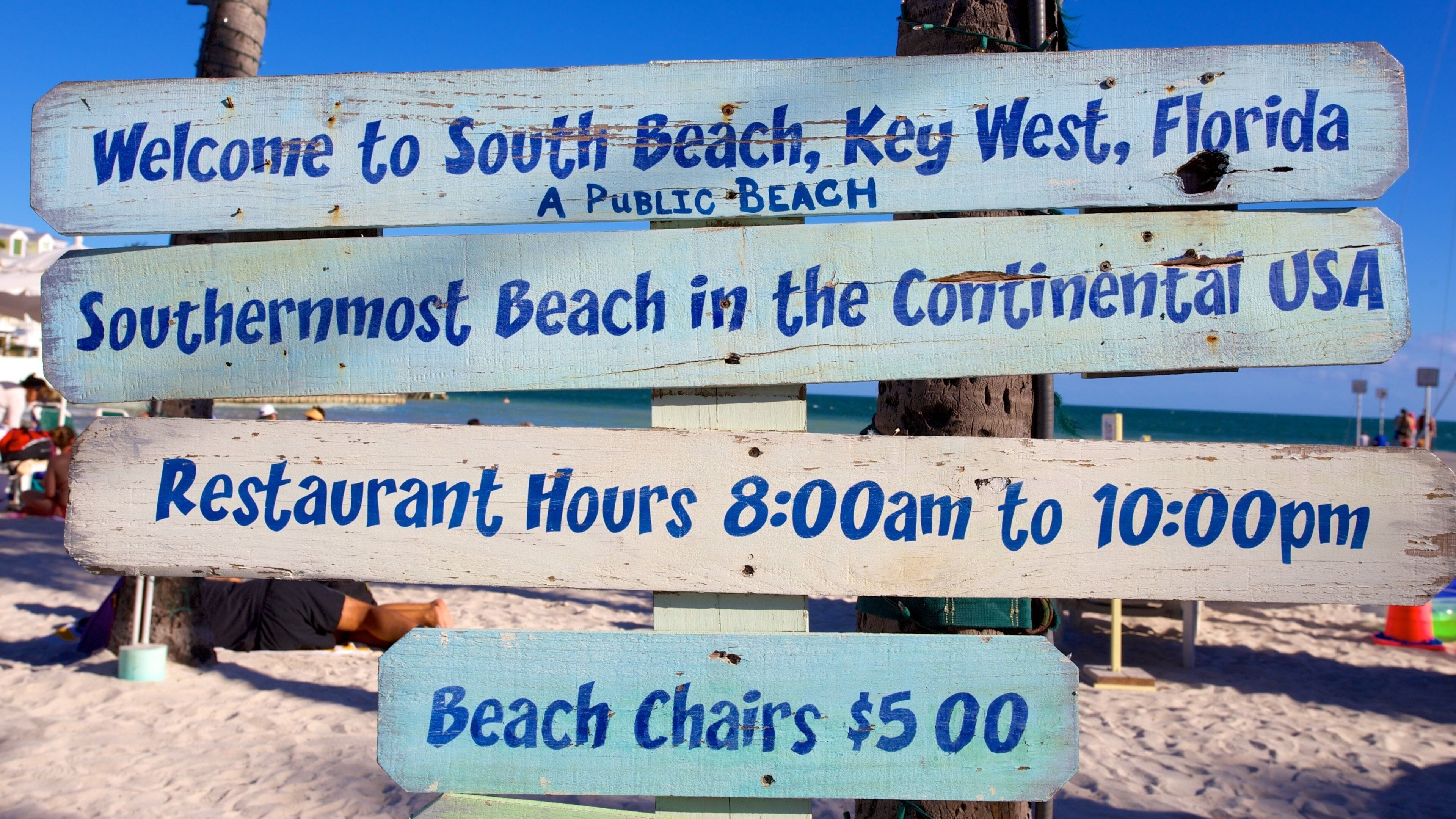 A wooden sign with information about South Beach, Key West, Florida, including public beach status, restaurant hours, and beach chair rental.