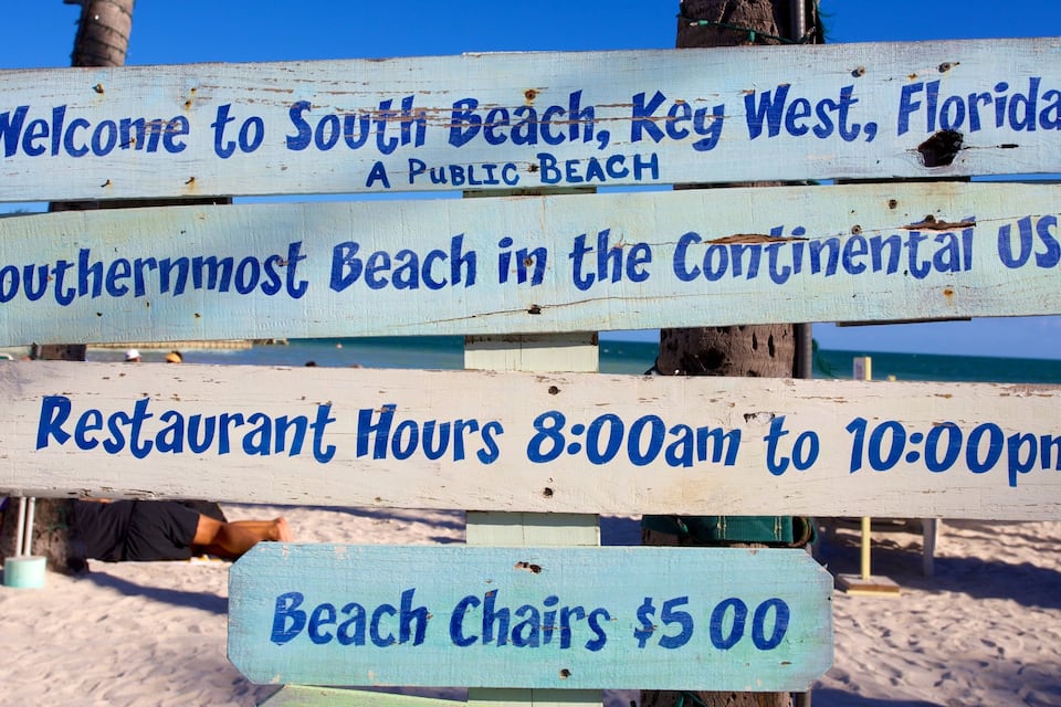A wooden sign with information about South Beach, Key West, Florida, including public beach status, restaurant hours, and beach chair rental.