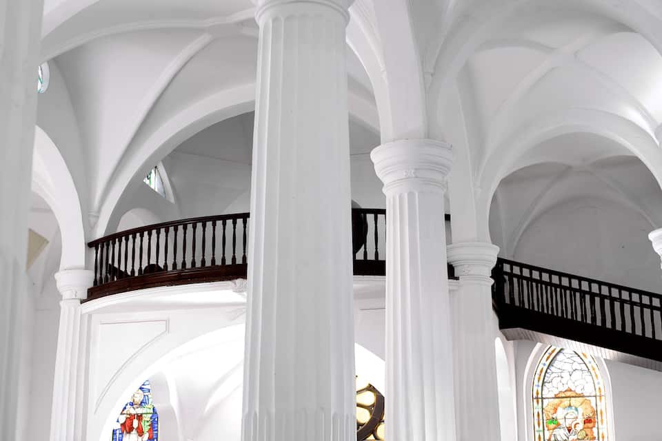 Interior de una iglesia con columnas blancas, un balcón de madera y vidrieras.