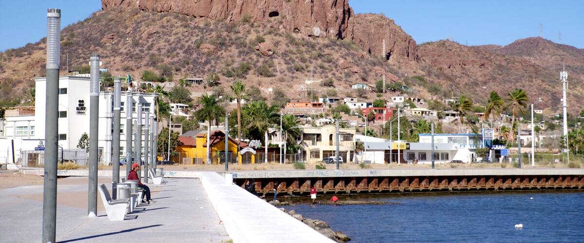 A coastal city with a prominent rocky mountain in the background, a waterfront promenade, and residential buildings.