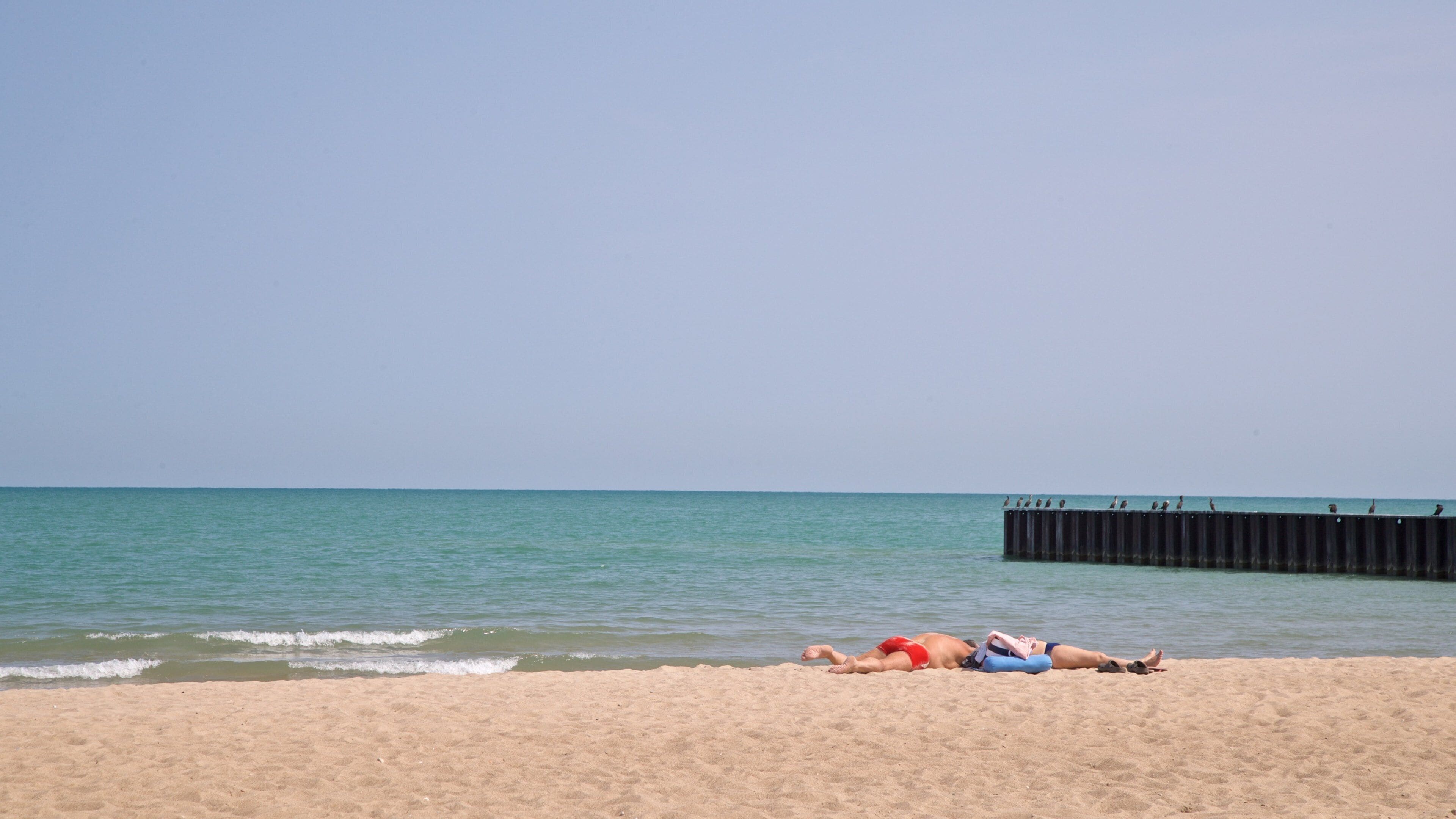 A beach with two people sunbathing, a wooden pier, and calm sea.